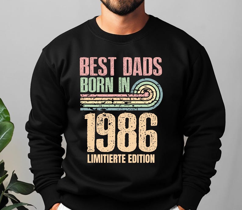 Op de afbeelding: Een zwart sweatshirt met lange mouwen en een grafische print met de tekst BEST DADS BORN IN 1986 LIMITIERTE EDITION. Het ontwerp bevat een verweerde, meerkleurige zonsondergangboog in roze, groen en blauw boven het grote jaartal 1986.