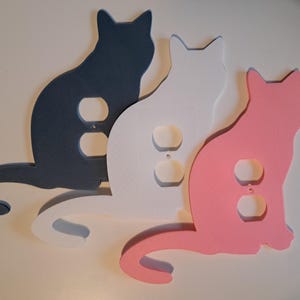 May include: Three cat-shaped outlet covers in navy blue, white, and pink. Each cat has two outlet holes. The cats are arranged side by side, with their tails curving to the right.