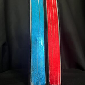 May include: Two hardcover books with black spines. One book has blue-edged pages, the other has red-edged pages. The pages are visible and create a colorful contrast against the black covers. The books are standing upright.