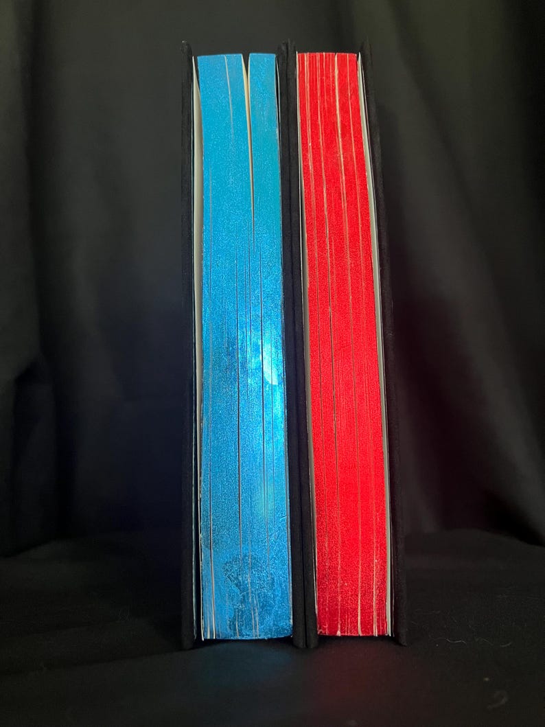 May include: Two hardcover books with black spines. One book has blue-edged pages, the other has red-edged pages. The pages are visible and create a colorful contrast against the black covers. The books are standing upright.