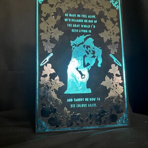May include: A black and teal illuminated art piece with a romantic theme. The artwork features a silhouette of a couple embracing, surrounded by floral designs and text that reads "He made me feel alive...and taught me how to see colors again."