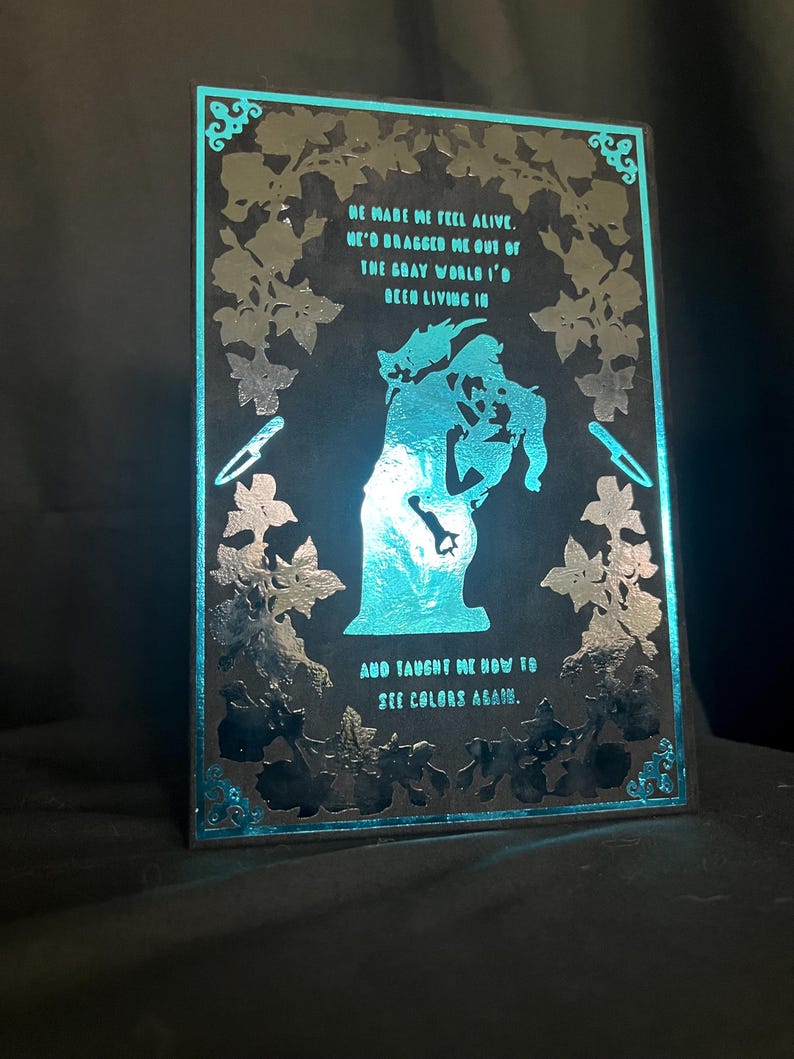 May include: A black and teal illuminated art piece with a romantic theme. The artwork features a silhouette of a couple embracing, surrounded by floral designs and text that reads "He made me feel alive...and taught me how to see colors again."