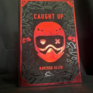 May include: A book cover with the title "CAUGHT UP" in red, featuring a red helmet design with a heart and cross symbol. The author's name, "NAVESSA ALLEN," is below. The cover has a black background with red accents.