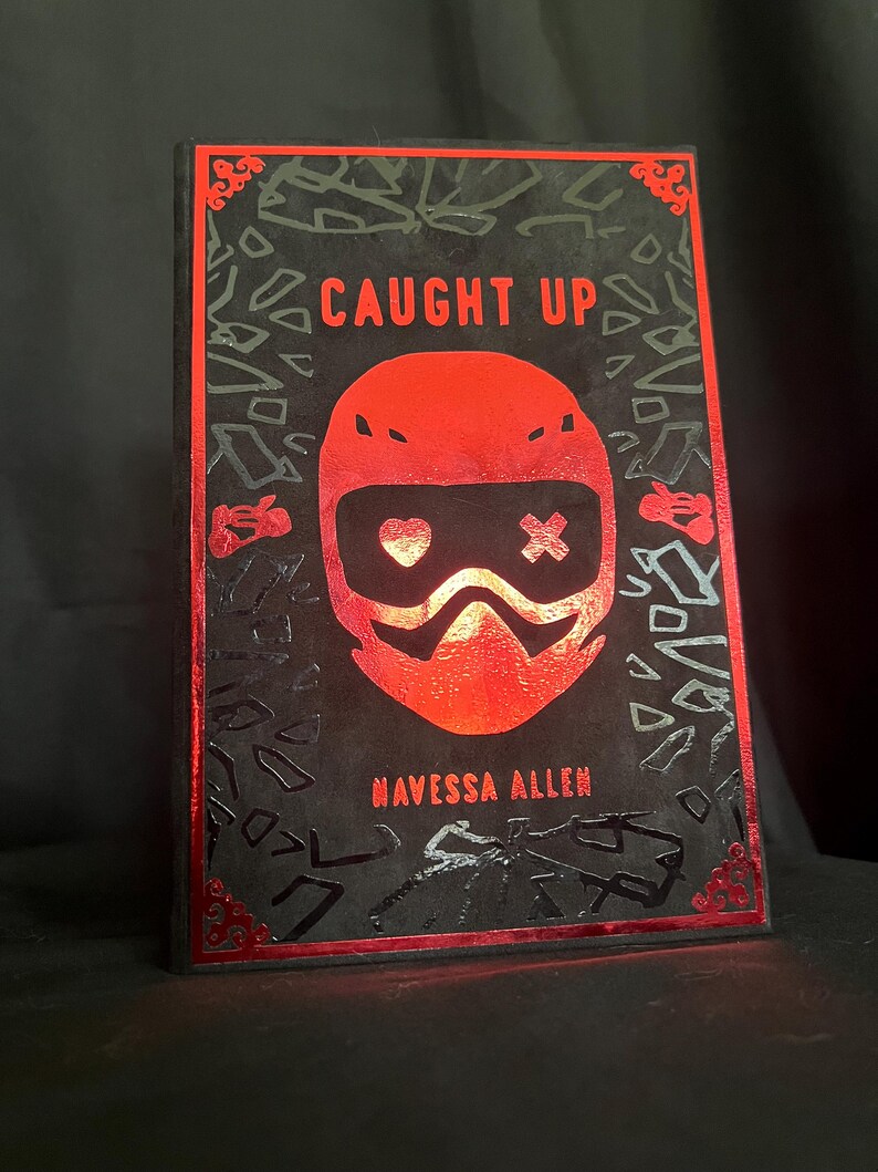 May include: A book cover with the title "CAUGHT UP" in red, featuring a red helmet design with a heart and cross symbol. The author's name, "NAVESSA ALLEN," is below. The cover has a black background with red accents.