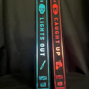 May include: Two hardcover books with black spines. The left book has a teal spine with a skull and knife graphic, the title "LIGHTS OUT", and the author's name. The right book has a red spine with a skull graphic, the title "CAUGHT UP", and the author's name.