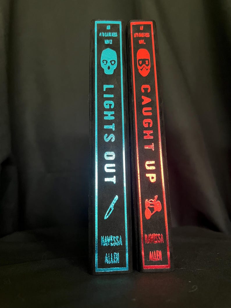 May include: Two hardcover books with black spines. The left book has a teal spine with a skull and knife graphic, the title "LIGHTS OUT", and the author's name. The right book has a red spine with a skull graphic, the title "CAUGHT UP", and the author's name.