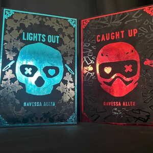 May include: Two framed art prints. One features a teal skull with the text "LIGHTS OUT" and the author's name. The other features a red helmet with the text "CAUGHT UP" and the author's name. Both are on a black background.