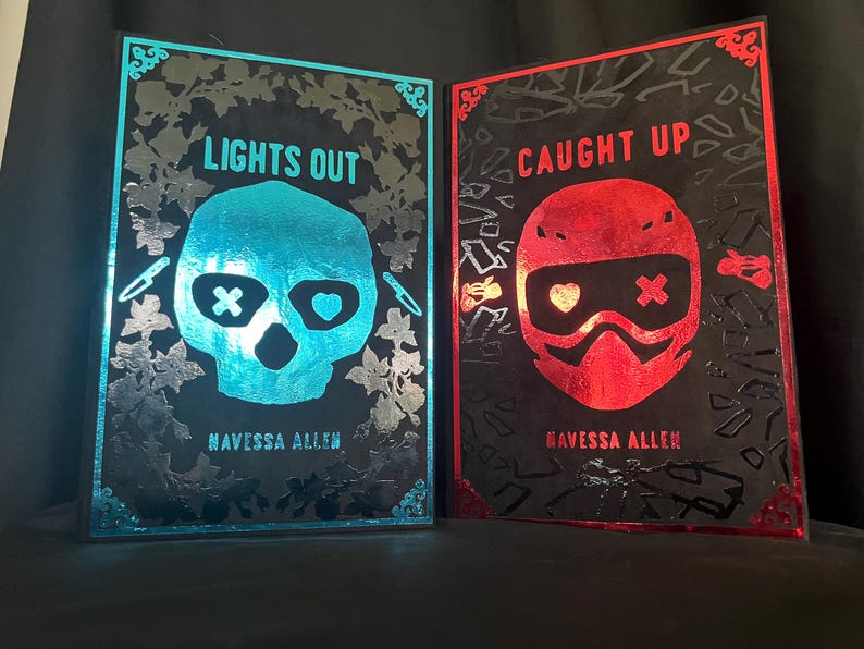 May include: Two framed art prints. One features a teal skull with the text "LIGHTS OUT" and the author's name. The other features a red helmet with the text "CAUGHT UP" and the author's name. Both are on a black background.