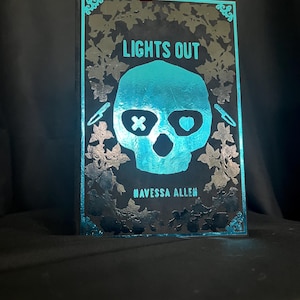 May include: A black and teal decorative book with the title "LIGHTS OUT" and the author's name, "NAVESSA ALLEN." The cover features a teal skull with an X for an eye and a heart for the other eye. The book is surrounded by a floral design.