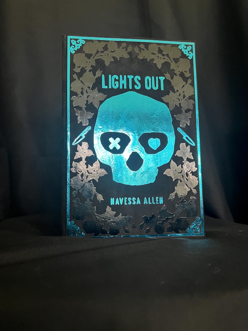 May include: A black and teal decorative book with the title "LIGHTS OUT" and the author's name, "NAVESSA ALLEN." The cover features a teal skull with an X for an eye and a heart for the other eye. The book is surrounded by a floral design.