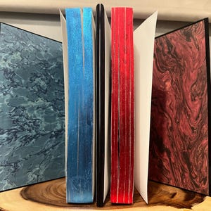 May include: A collection of handmade journals with marbled covers in shades of blue, red, and burgundy. The edges of the pages are colored blue and red. The journals are displayed open, revealing the interior pages.