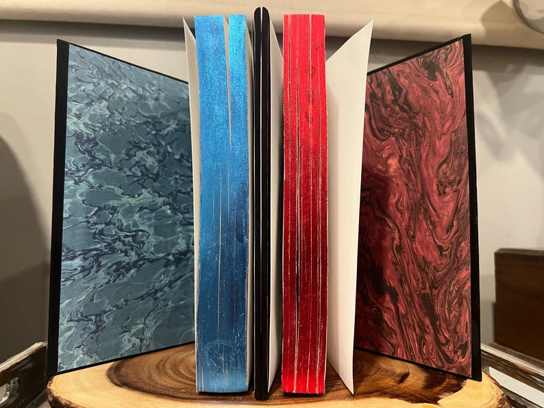 May include: A collection of handmade journals with marbled covers in shades of blue, red, and burgundy. The edges of the pages are colored blue and red. The journals are displayed open, revealing the interior pages.