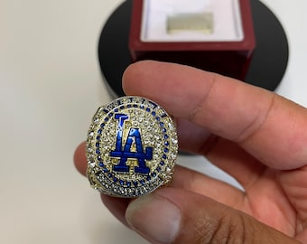 May include: A gold-colored championship ring with a blue "LA" logo, surrounded by blue and clear gemstones. The ring is held in a hand, with a red ring box in the background.
