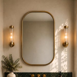 Op de afbeelding: Een badkamermeubel met een donkergroen marmeren blad met witte aders, een gouden kraan en een geribbelde houten onderkast. Erboven hangt een ovale spiegel met een gouden lijst, geflankeerd door twee cilindervormige glazen wandlampen.
