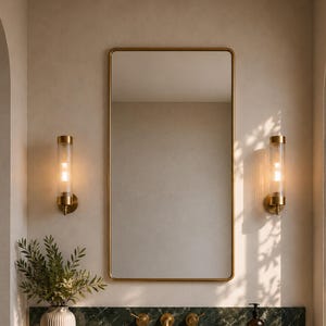 May include: A bathroom vanity featuring a dark green marble countertop with a gold faucet and handles. Above, a rectangular mirror with a gold frame is flanked by two cylindrical glass wall sconces with gold hardware. A ribbed wooden cabinet base.