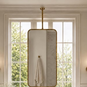 May include: A rectangular mirror with rounded corners and a metallic brass frame, suspended from the ceiling by a vertical brass rod. The mirror reflects a towel hanging on a hook and a marble countertop with a sink, faucet, and accessories.