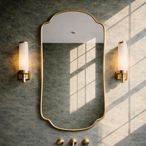 Puede incluir: Un mueble de baño con un espejo con marco dorado, dos apliques y un lavabo de piedra. El espejo tiene una forma única y los apliques tienen pantallas cilíndricas. El lavabo tiene un diseño minimalista.