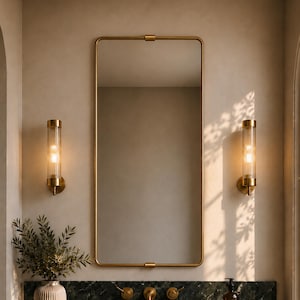 May include: A rectangular wall mirror with a thin brass frame centered above a dark green marble vanity countertop. Two cylindrical glass wall sconces with brass accents flank the mirror. A ribbed vase with greenery, folded towels, and a soap dispenser sit on the counter.