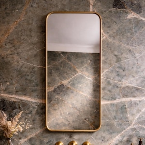 May include: A rectangular gold-framed mirror hangs above a marble sink with gold fixtures. The mirror reflects a glimpse of a white ceiling. A tray with a vase of dried flowers and bottles sits on the counter, complementing the luxurious bathroom design.