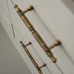 Peut inclure: Gros plan sur deux poignées de placard en laiton antique sur une porte de placard gris clair. Les poignées présentent un design détaillé et texturé et une finition chaude et vieillie. La porte de l'armoire a un design de panneau encastré.