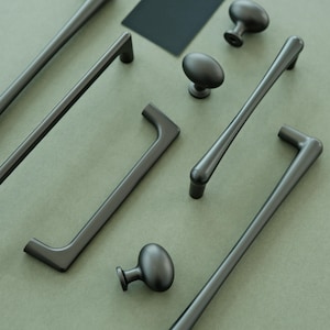 May include: A collection of black cabinet hardware, including pulls and knobs, arranged on a green surface. The hardware is made of metal and has a matte finish.