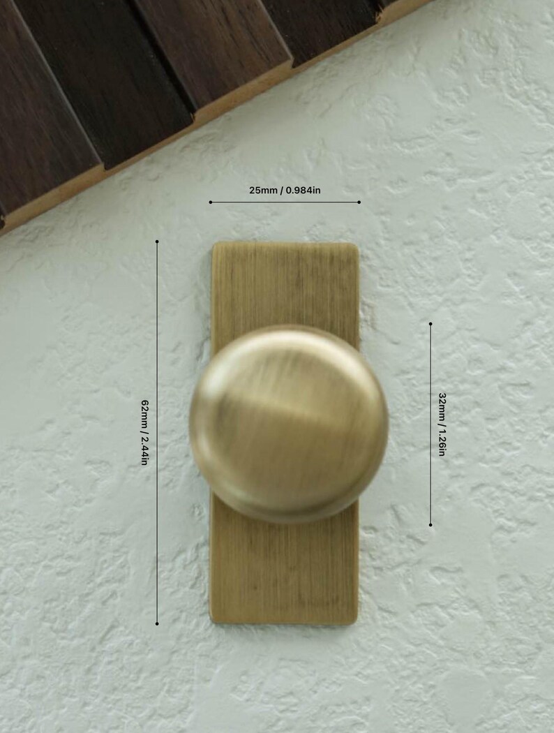 May include: A brass cabinet knob with a rectangular backplate and a rounded knob. The knob measures 3.2 cm in height and 6.2 cm in length. The backplate is 2.5 cm wide. The hardware is mounted on a textured, off-white surface.
