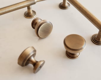 May include: An assortment of antique brass cabinet hardware, including bar pulls and round knobs. The hardware has a warm, aged finish and a classic design, suitable for kitchen or bathroom cabinetry. The hardware is displayed on a light-colored surface.