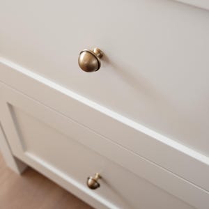 May include: Close-up of a white cabinet with two drawers. Each drawer has a round, antique brass knob. The knobs are centered on the drawers. The cabinet is set against a light-coloured wooden floor.