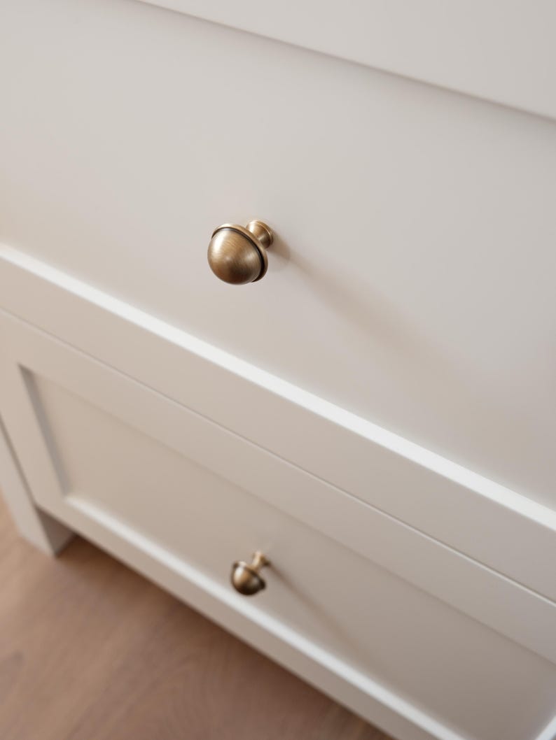 May include: Close-up of a white cabinet with two drawers. Each drawer has a round, antique brass knob. The knobs are centered on the drawers. The cabinet is set against a light-coloured wooden floor.