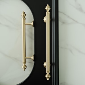 May include: Two gold-colored door handles with ornate finials and a matte finish. The handles are mounted on a black door frame with a white marble-patterned background. The handles are approximately 12 inches in length.