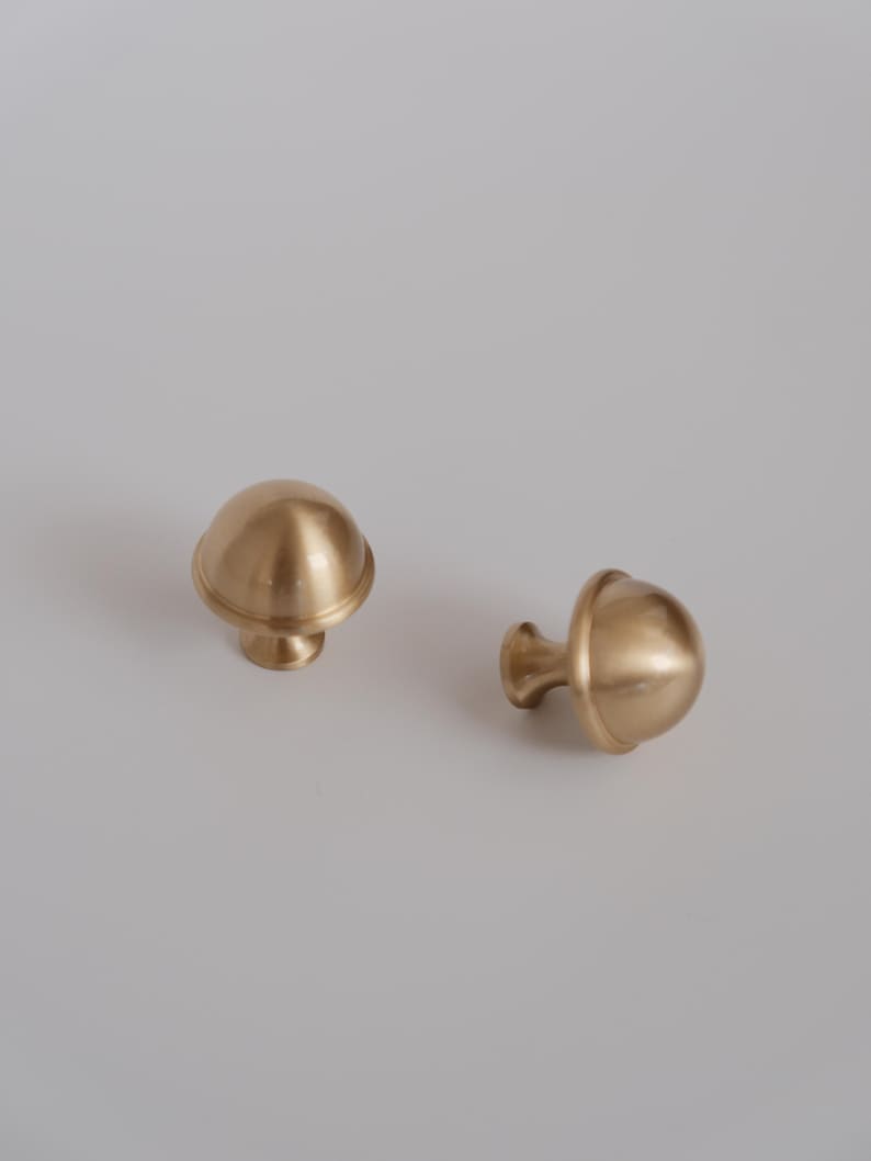 May include: Two brass cabinet knobs with a mushroom-shaped design. The knobs have a rounded top and a short, cylindrical base. The finish is a warm, golden colour. The knobs are likely for furniture or cabinetry.