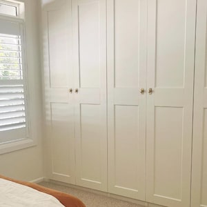 May include: A series of white, panelled wardrobe doors with gold-coloured handles. The doors are in a bedroom with neutral carpeting and a bed with a rust-coloured blanket. A window with white blinds is visible on the left.