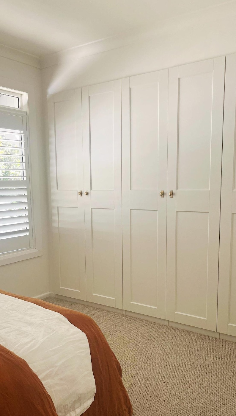 May include: A series of white, panelled wardrobe doors with gold-coloured handles. The doors are in a bedroom with neutral carpeting and a bed with a rust-coloured blanket. A window with white blinds is visible on the left.