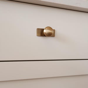 May include: Close-up of a white cabinet drawer with a brushed brass knob. The knob has a circular design and is mounted on a rectangular backplate. The cabinet is part of a larger piece of furniture.