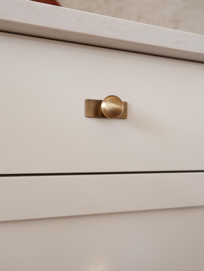 May include: Close-up of a white cabinet drawer with a brushed brass knob. The knob has a circular design and is mounted on a rectangular backplate. The cabinet is part of a larger piece of furniture.