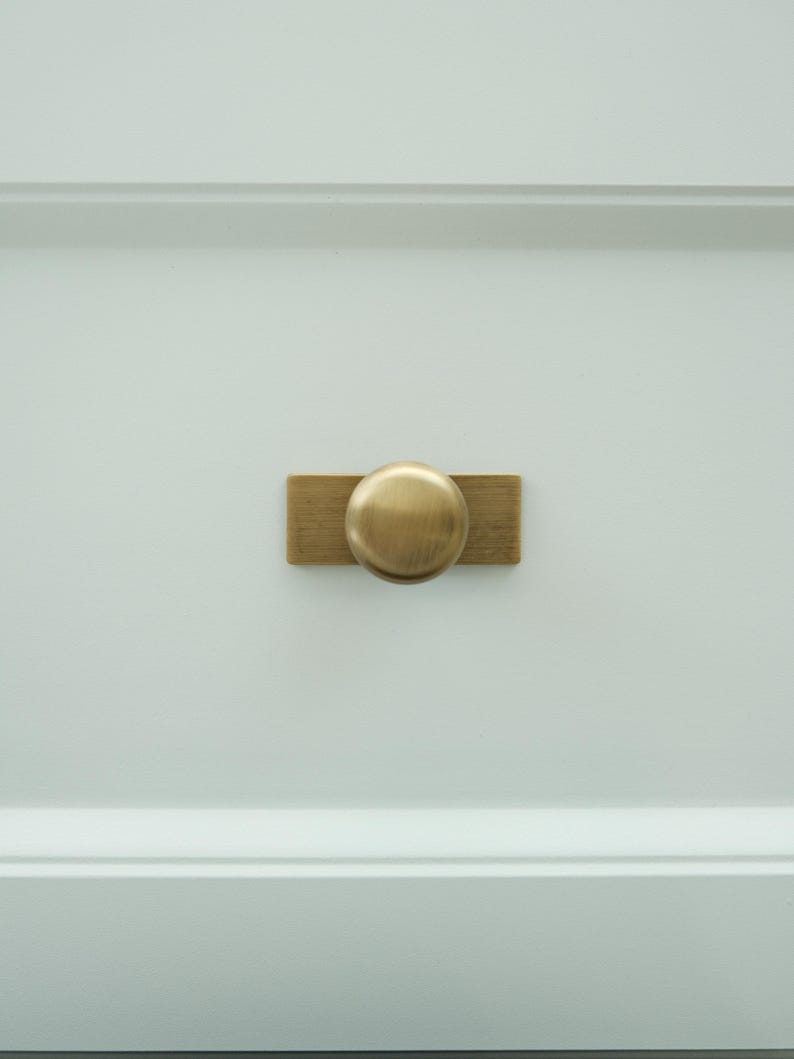 May include: A close-up of a white cabinet door with a brass knob and rectangular backplate. The knob is round and the backplate is rectangular. The hardware is centered on the door.