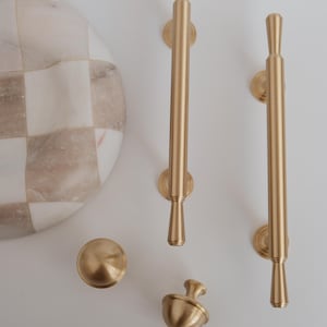 May include: A selection of gold-coloured cabinet hardware, featuring two long pulls, a round knob, and an acorn-shaped knob. A marble and beige checkered serving board is also visible.
