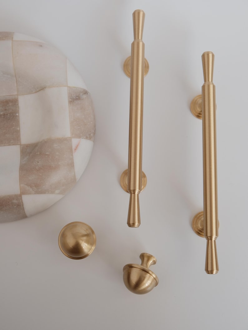 May include: A selection of gold-coloured cabinet hardware, featuring two long pulls, a round knob, and an acorn-shaped knob. A marble and beige checkered serving board is also visible.