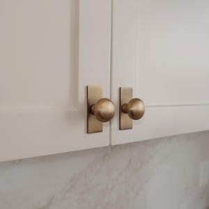 Puede incluir: Primer plano de dos puertas de armario blancas con herrajes de color latón. Cada puerta tiene una placa trasera rectangular y un pomo redondo. Los herrajes tienen un acabado metálico cálido, que contrasta con los armarios blancos. El fondo muestra una superficie similar al mármol.