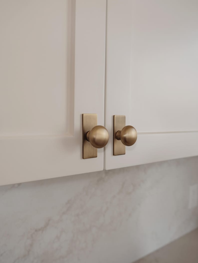 May include: Close-up of two white cabinet doors with brass-coloured hardware. Each door has a rectangular backplate and a round knob. The hardware has a warm, metallic finish, contrasting with the white cabinets. The background shows a marble-like surface.