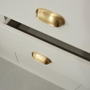 May include: Close-up of a white cabinet with two brass-coloured, cup-style drawer pulls. The pulls are curved and have a polished finish. The cabinet appears to be new and modern, with clean lines and a minimalist design.