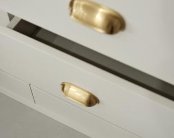May include: Close-up of a white cabinet with two brass-colored, cup-style drawer pulls. The pulls are curved and have a polished finish. The cabinet appears to be new and modern, with clean lines and a minimalist design.