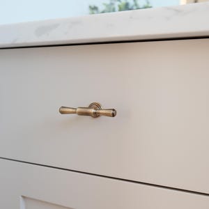 Könnte beinhalten: Nahaufnahme einer hellgrauen Schublade mit einem messingfarbenen Griff. Der Griff ist ein horizontaler Stab mit abgerundeten Enden. Die Arbeitsplatte darüber ist weiß mit grauer Maserung. Der Griff ist etwa 10 cm lang.