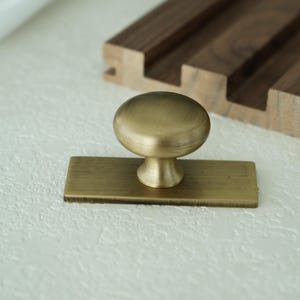 May include: A brass cabinet knob with a rounded top and a rectangular base. The knob is set against a textured white surface, with a wooden accent in the background.
