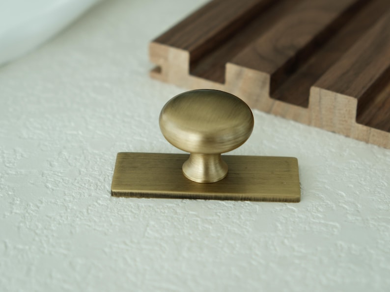 May include: A brass cabinet knob with a rounded top and a rectangular base. The knob is set against a textured white surface, with a wooden accent in the background.