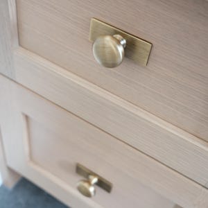 May include: Close-up of two wooden cabinet drawers with brass hardware. Each drawer has a round knob on a rectangular backplate. The wood has a light, natural finish, and the hardware has a warm, antique brass tone.