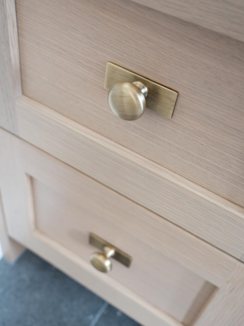 May include: Close-up of two wooden cabinet drawers with brass hardware. Each drawer has a round knob on a rectangular backplate. The wood has a light, natural finish, and the hardware has a warm, antique brass tone.
