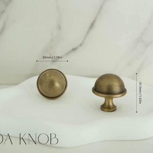 May include: Two antique brass ADA KNOB cabinet knobs. One knob is round, 30mm in diameter, and the other is a mushroom shape, 31.1mm tall. Both knobs are displayed on a white, cloud-shaped tray.