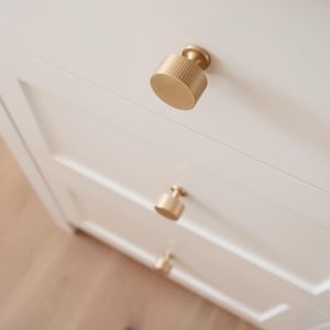 Könnte beinhalten: Nahaufnahme eines weißen Schranks mit drei Schubladen, die jeweils mit einem zylindrischen, goldfarbenen Schrankknauf versehen sind. Die Knöpfe haben eine geriffelte Textur und eine kreisförmige Basis. Der Schrank steht auf einem hellen Holzboden.