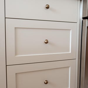 May include: Close-up of three off-white kitchen cabinet drawers with brass-coloured knobs. The drawers are rectangular with a recessed panel design. The top drawer is plain, while the bottom two have a framed appearance. The cabinets are part of a larger kitchen setup.